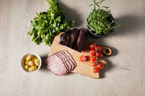 top view of of tasty ham on cutting board with greenery, cherry tomatoes, olives on wooden table