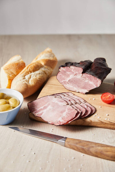 selective focus of tasty ham on cutting board with knife, cherry tomato, olives and baguette on wooden table isolated on grey