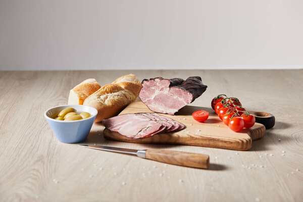 selective focus of tasty ham on cutting board with knife, cherry tomatoes, olives and baguette on wooden table isolated on grey