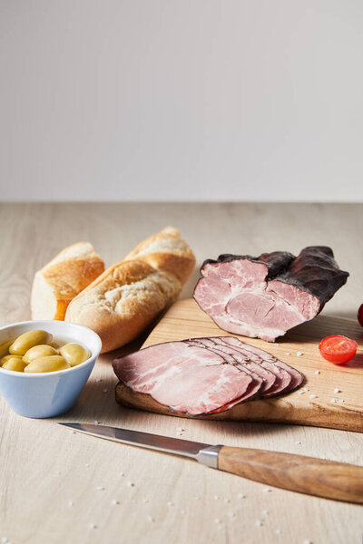 tasty ham on cutting board with knife, cherry tomato, olives and baguette on wooden table isolated on grey