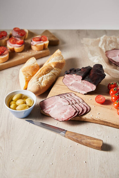 selective focus of tasty ham on cutting board with knife, cherry tomatoes, olives and baguette near canape on wooden table isolated on grey