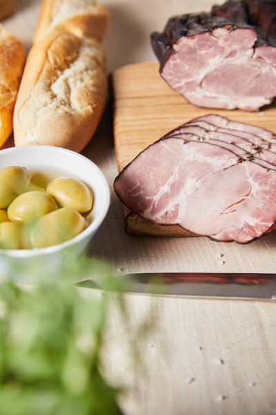 selective focus of tasty ham on cutting board with knife, olives and baguette on wooden table