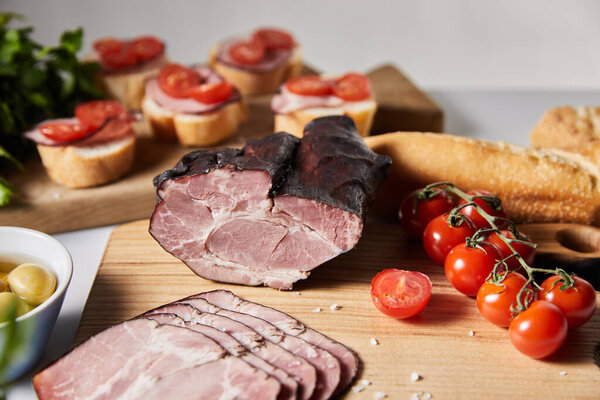 selective focus of tasty ham on cutting board with parsley, cherry tomatoes, olives and baguette near canape isolated on grey