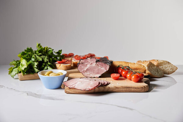 tasty ham on cutting board with parsley, cherry tomatoes, olives and baguette on white marble surface isolated on grey