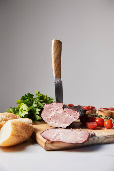 selective focus of knife in tasty ham on cutting board with parsley, cherry tomatoes and baguette on white surface isolated on grey
