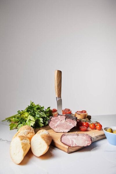 selective focus of knife in tasty ham on cutting board with parsley, cherry tomatoes and baguette on white surface isolated on grey