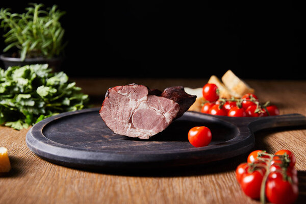 selective focus of tasty ham on board near parsley, cherry tomatoes and baguette on wooden table isolated on black