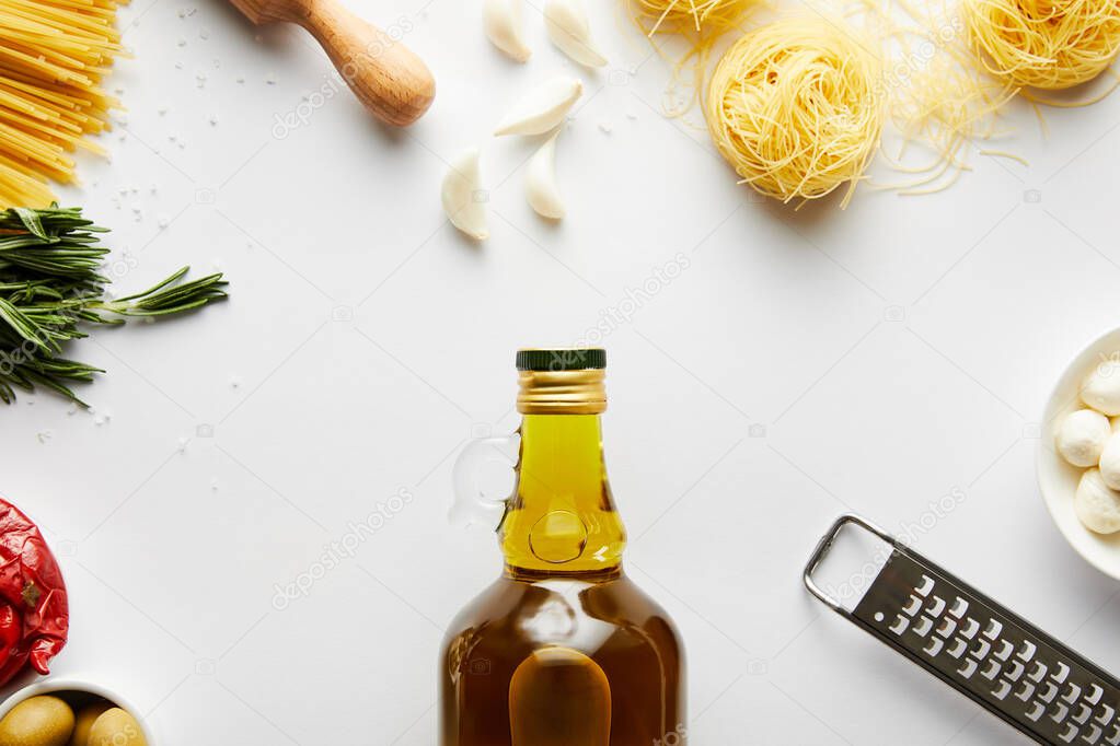 Top view of bottle of olive oil, rolling pin, grater, pasta and ingredients on white 