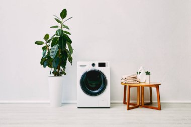 Modern çamaşır makinesinin yanındaki yeşil bitki havluları ve banyoda deterjan şişesi olan kahve masası. 