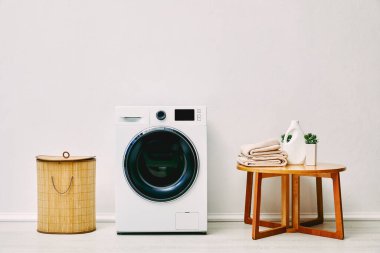 Çamaşır sepetinin yanında modern çamaşır makinesi, havluyla kahve masası, deterjan şişesi ve banyo bitkisi. 