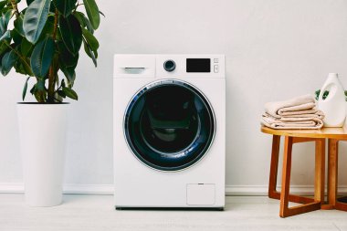 Çamaşır makinesinin yanındaki yeşil bitki, banyoda havlu ve deterjan şişesi olan ahşap sehpa. 