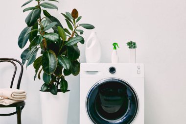Modern çamaşır makinesinin yanında yeşil bitkiler ve banyoda havluları olan sandalyelerin yanında şişeler. 