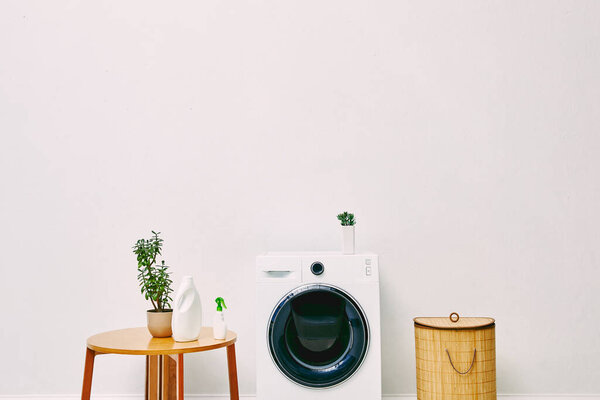 green plants and bottles near wooden coffee table, laundry basket and modern washing machine in bathroom 