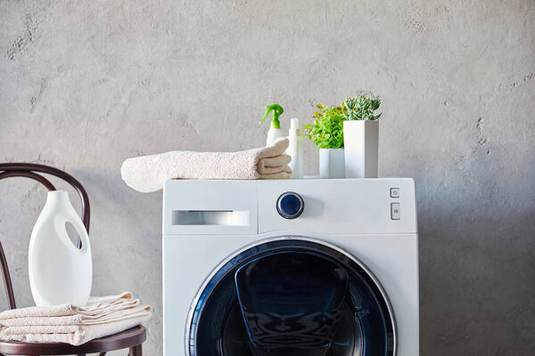 detergent and spray bottles on washing machine near plants, towels and chair in bathroom 