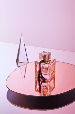 Kristal şeffaf piramit. Pembe arka planda, yuvarlak aynada parfüm şişesinin yanında yansıması var.