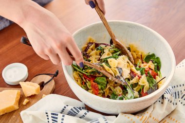 Makarna salatasını peçetenin yanında spatulayla karıştıran bir kadının kısmi görüntüsü, mutfak masasının yanında parmesanla tuz ve kesim tahtası.