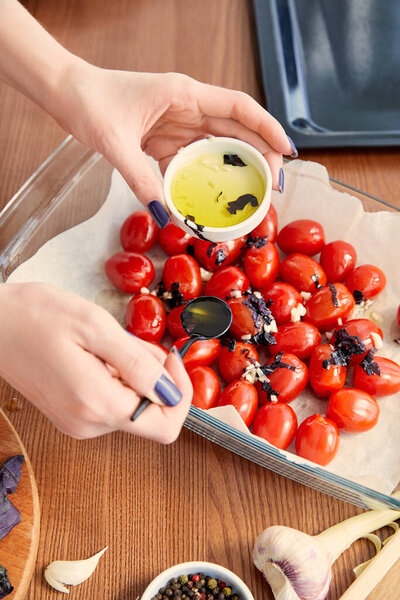 Cropped view of woman adding olive oil to tomatoes with ingredients near oven tray on wooden background