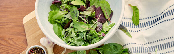 Horizontal image of napkin and bowls with pepper and lettuce with herbs near garlic on cutting board on wooden background