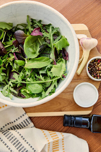 Top view of garlic and bowl with ingredients on cutting board near napkin and bottle of olive oil on wooden background