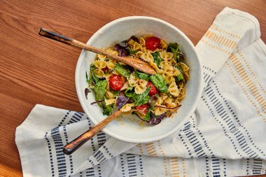 Tahta arka planda peçetenin yanında makarna salatası ve spatula bulunan kasenin en üst görüntüsü.