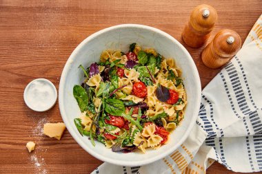 Makarna salatası, peçete, parmesan, biber ve ahşap arka planda tuz değirmenlerinin olduğu kasenin üst görüntüsü.
