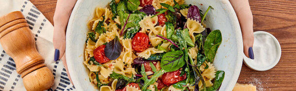 Cropped view of woman holding bowl with pasta salad near napkin and salt mill on wooden background, panoramic shot