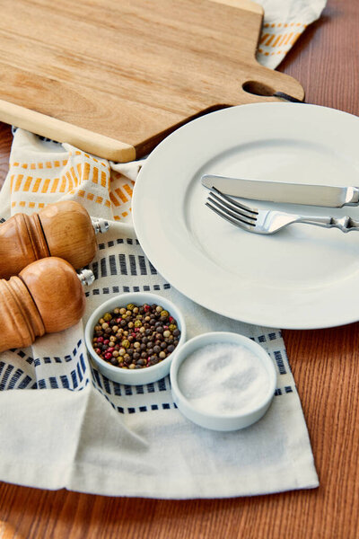 Высокий угол обзора пластины с ножом и вилкой рядом с тканью, разделочной доской, чашами, перцовыми и солеными мельницами на деревянном фоне
