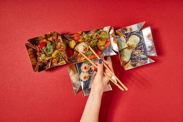 top view of woman holding chopsticks with steamed bun near prepared chinese food in takeaway boxes on red