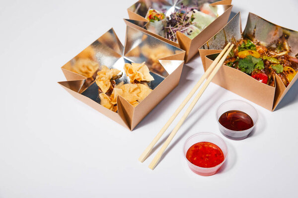 sauces and chopsticks near takeaway boxes with prepared chinese food on white 