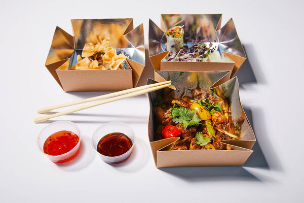 spicy sauces and chopsticks near takeaway boxes with prepared chinese food on white 