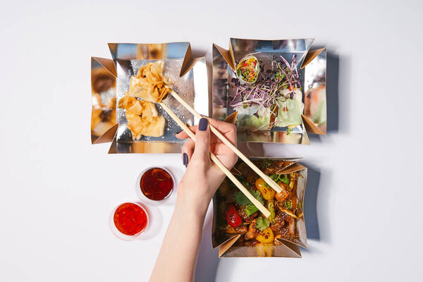 top view of woman holding chopsticks near takeaway boxes with prepared chinese food and sauces on white 