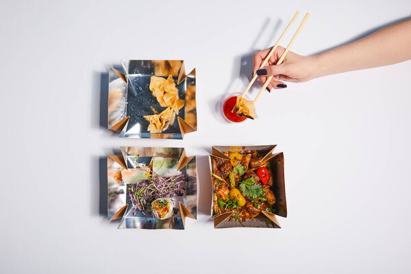 top view of woman holding chopsticks with dumpling near sauce and takeaway boxes with prepared chinese food on white 