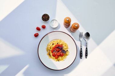 top view of delicious pasta with tomatoes served with cutlery, salt and pepper mills on white table in sunlight clipart