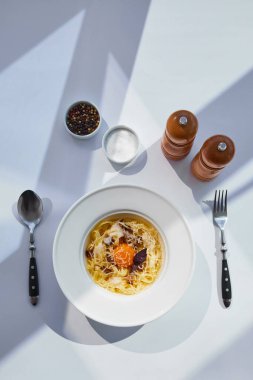 Güneş ışığında beyaz masa üzerinde çatal bıçak, tuz ve biber değirmenleriyle servis edilen lezzetli carbonara makarna manzarası