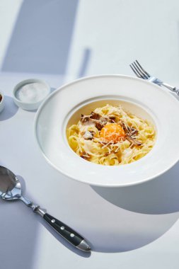 Carbonara makarnası, çatal bıçak takımı ve tuzlu güneş ışığı altında beyaz masada servis edilir.