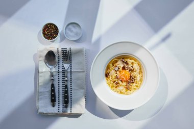 Çatal bıçakla servis edilen lezzetli carbonara makarnasının üst görüntüsü ve güneş ışığında beyaz masada baharat.