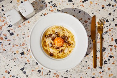 top view of delicious pasta carbonara served with golden cutlery, salt and pepper shakers on stone table in sunlight clipart