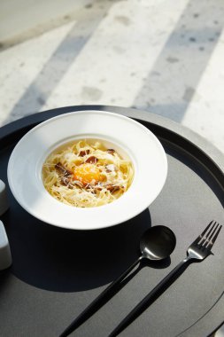 Lezzetli makarna carbonara ile servis edilir siyah masa üzerinde çatal bıçak mermer arka planda güneş ışığı