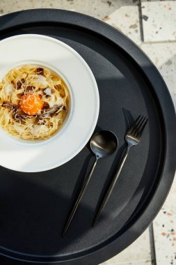 Nefis Carbonara makarnasının üst görüntüsü. Güneş ışığında siyah masada servis edilen çatal bıçak seti.