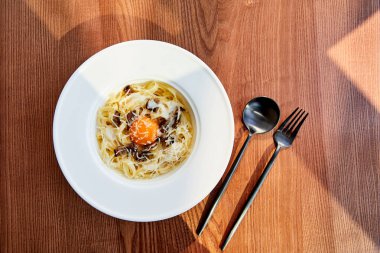 top view of delicious pasta carbonara served with black cutlery on wooden background in sunlight clipart