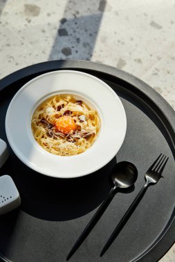 Lezzetli makarna carbonara ile servis edilir siyah masa üzerinde çatal bıçak mermer arka planda güneş ışığı