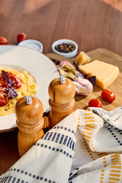 selective focus of delicious pasta with tomatoes served on wooden table with cutlery, napkin, seasoning and ingredients in sunlight