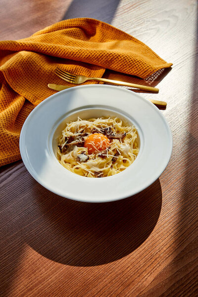 delicious pasta carbonara served with golden cutlery and yellow napkin on wooden table in sunlight