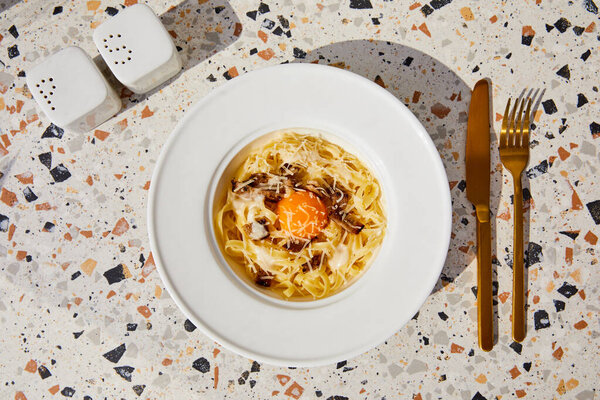 top view of delicious pasta carbonara served with golden cutlery, salt and pepper shakers on stone table in sunlight