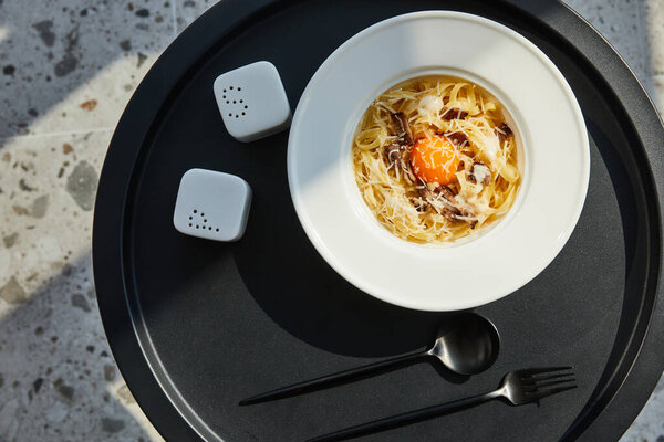 top view of delicious pasta carbonara served with cutlery, seasoning on black table on marble background in sunlight
