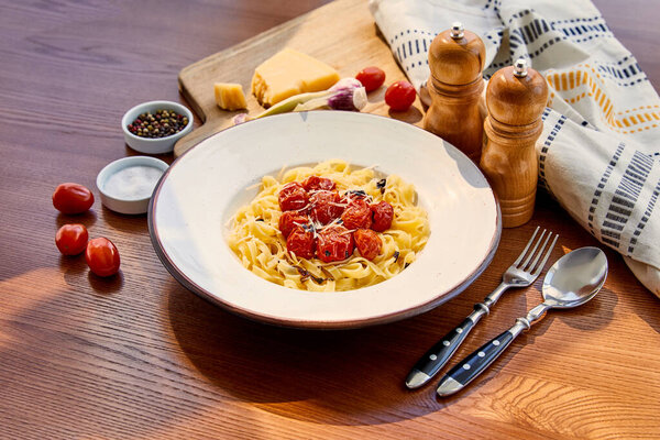 delicious pasta with tomatoes served on wooden table with cutlery, napkin, seasoning and ingredients in sunlight