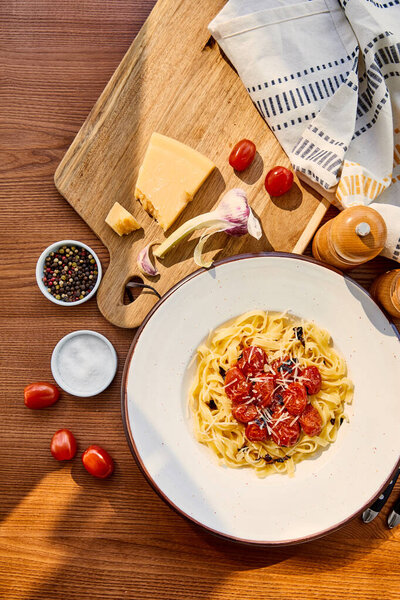 top view of delicious pasta with tomatoes served on wooden table with cutlery, napkin, seasoning and ingredients in sunlight