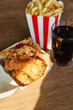 selective focus of deep fried chicken, french fries and soda in glass on wooden table in sunlight clipart