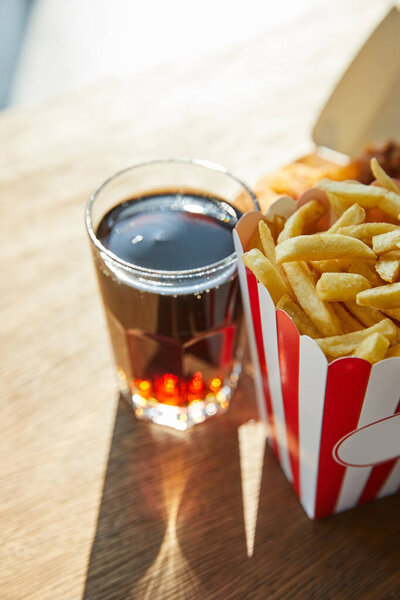 selective focus of french fries and soda in glass on wooden table in sunlight