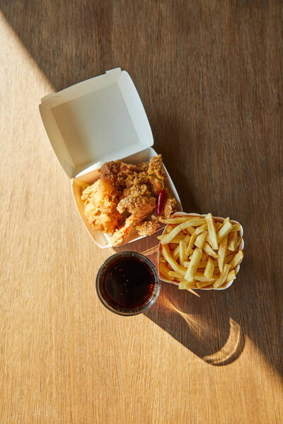 top view of deep fried chicken, french fries and soda in glass on wooden table in sunlight